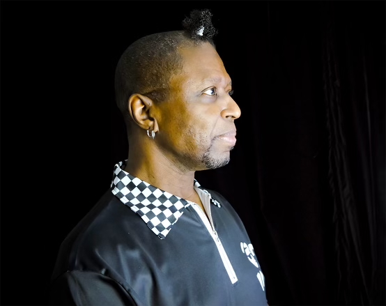 DJ Foxxy, a black man, is seen in profile fron the shoulders up. His hair is cropped very short except for a perfect little ponytail at the top of his forehead. He's wearing a black shirt with white piping an a checkerboard collar. He's holding back a natural smile in order to look serious.